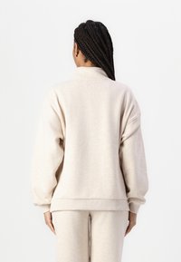 Beige színű pulóver magas nyakkal, esett vállakkal és bordás mandzsettákkal. Sima textúrájú, laza szabású, azonos színű alsóval párosítva.