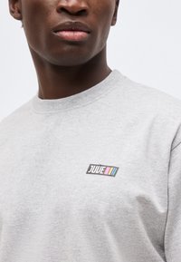 Κοντινό πλάνο ενός ατόμου που φορά ένα ανοιχτό γκρι φούτερ με στρογγυλή λαιμόκοψη και ένα μικρό πολύχρωμο λογότυπο "JUVE" στο στήθος.