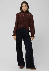 Blouse en dentelle bourgogne à col montant et poignets à volants, associée à un pantalon large en velours côtelé bleu marine et des chaussures à plateforme marron.