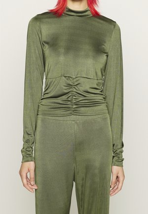 Personne aux cheveux roux portant un haut froncé à manches longues vert olive et un pantalon assorti sur un fond clair uni.