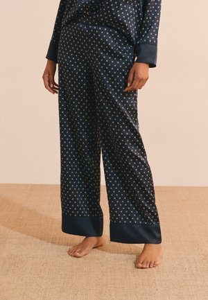Personne portant un pantalon de pyjama en satin bleu marine avec de petits motifs de croix dorées, debout pieds nus sur un sol beige texturé.