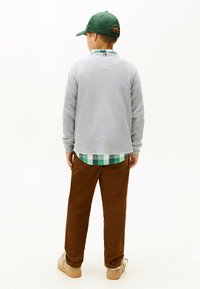 Maglione a rete grigio, camicia a quadri verde sotto, pantaloni marroni e scarpe beige. Berretto da baseball verde in testa. Vista posteriore.