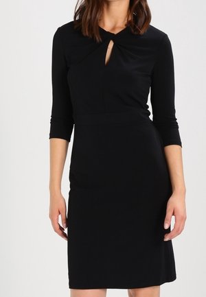 Femme portant une robe noire ajustée avec des manches trois-quarts et un petit détail en forme d'œillet à l'encolure, sur un fond uni.