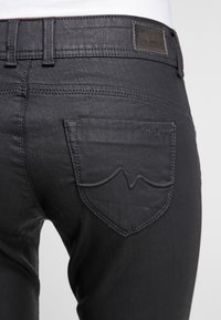 Svarta denimjeans har ett strukturerat tyg, en bakficka med ett distinkt kurvigt sömnadsmönster, och en läderloggopatch på midjebandet.