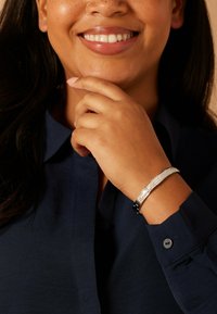 Zilveren armband met een gestructureerd ontwerp, met heldere accenten en een zwarte sluiting. Draagt op een hand die rust bij de kin, tegen een blauw shirt.