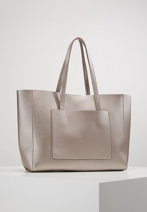 Große metallische beige Tragetasche mit zwei Griffen und einer äußeren Vordertasche, auf einem weißen Sockel vor einfachem Hintergrund platziert.