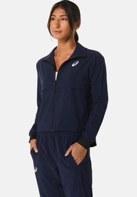 Frau in einem marineblauen Asics-Trainingsanzug mit einer Reißverschlussjacke und Hose, steht mit den Händen in den Taschen vor einem einfarbigen Hintergrund.