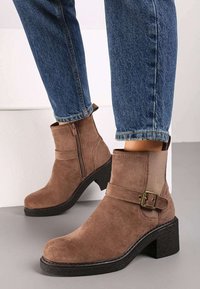Bottines en daim marron avec fermeture éclair et accent de boucle, présentant un talon noir épais et une texture lisse. Portées avec un jean bleu.