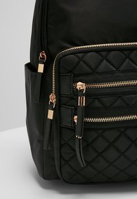 Poche de sac à dos matelassé noir avec fermetures éclair dorées et tirettes à pompons noirs, sur fond gris et blanc.