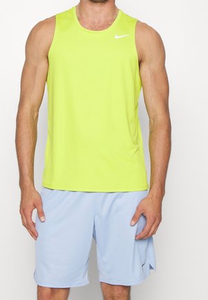 Mężczyzna w neonowożółtej, bezrękawnikowej koszulce sportowej i jasnoniebieskich, sportowych szortach, stojący z ramionami swobodnie opuszczonymi wzdłuż ciała.