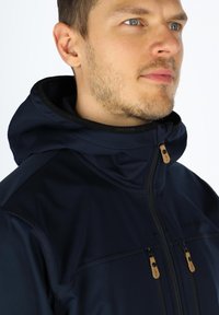 Marineblå jakke med hætte, der har kontrasterende lynlåse og beige detaljer. Glat tekstur med synlige sømme og logo på kraven.