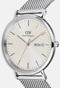 Hõbedane võrksuur kell, millel on tekstuurne kreemjas valge dial, hõbedased tunni numbrid ja kuupäeva näit, millel on kirjas "KOL 10". Brändi nimi "Daniel Wellington" on esile toodud.