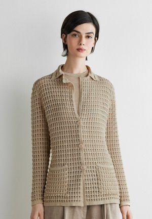 Kobieta ubrana w beżowy otwarty sweter z guzikami i dwoma przednimi kieszeniami na dopasowanym topie, stojąca na tle gładkiej białej ściany.