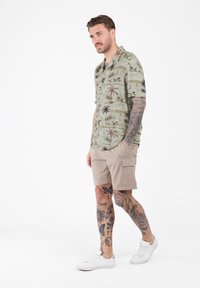 Hombre con tatuajes que lleva una camisa de manga corta con estampado tropical verde, pantalones cortos de carga beiges y zapatillas blancas, caminando frente a un fondo blanco.