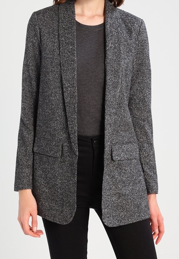 Blazer grigio testurizzato con vestibilità slim, revers a punta, tasche frontali e design aperto, indossato sopra una camicia scura e pantaloni neri.