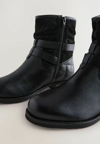 Botas de tobillo negras confeccionadas con materiales de cuero y ante, que presentan un cierre de cremallera, detalles de correas y una punta ligeramente afilada. Acabado texturizado.