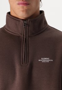 Brown, zipped mock neck sweatshirt made of soft fabric; features a logo in white text on the chest and prominent seams on the collar.