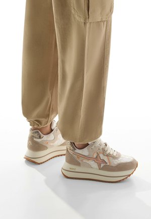 Sneaker beige con una toma superiore texturizzata, caratterizzato da una suola curva, dettagli arancioni e motivi decorativi. Abbinato a pantaloni beige a vestibilità larga.