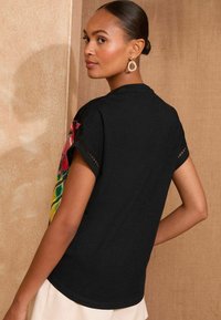 Must ja lühikeste varrukatega T-särk, millel on värvikas lilleline muster õlgadel, silmade detailidega varrukates ja ümmargune kaelus.