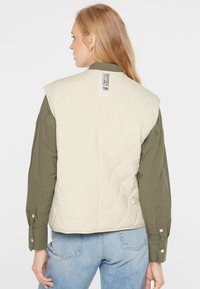 Frau mit langen blonden Haaren, die eine cremefarbene gesteppte Weste über einem olivgrünen Shirt und hellblauen Jeans trägt, von hinten gezeigt.