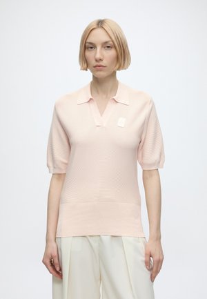 Femme aux cheveux blonds courts, portant un haut tricoté manches courtes rose pâle et un pantalon beige clair, debout devant un fond blanc uni.