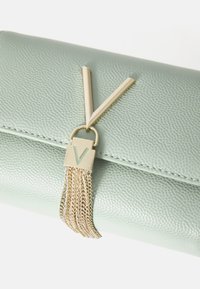 Clutch in pelle verde chiaro con logo 'V' color oro e dettaglio a frange realizzato con catene intrecciate. La superficie texturizzata conferisce profondità.