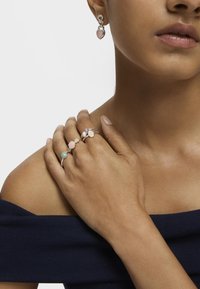 Pendientes de plata con acentos en forma de corazón y tres anillos con piedras de colores pastel en engastes circulares, llevados sobre un hombro desnudo.