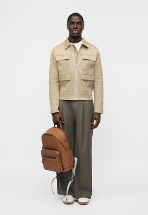 Man die een beige jas en grijze broek met wijd uitlopende pijpen draagt, en een bruine leren rugzak bij het handvat vasthoudt, staand tegen een effen achtergrond.