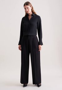 Zwarte blouse met lange mouwen en een kraag, gecombineerd met loszittende zwarte broek. Gladde stoftextuur en op maat gemaakt ontwerp.