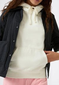 Czarna nylonowa kurtka na białej bluzie z kapturem, posiadającej sznurek. Bluza ma kieszeń kangurkę. Miękka tekstura, swobodny design i luźny krój.
