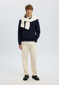 Suéter de punto azul marino con una bufanda color crema drapeada sobre los hombros, combinado con pantalones crema claros y zapatos negros. Diseño sencillo, tejido texturizado.