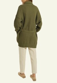 Frau, die einen übergroßen, olivgrünen Strickcardigan mit Gürtel, cremefarbene, schmal zulaufende Hosen und beigen Mokassin-Schuhen trägt, mit dem Rücken zur Kamera steht.