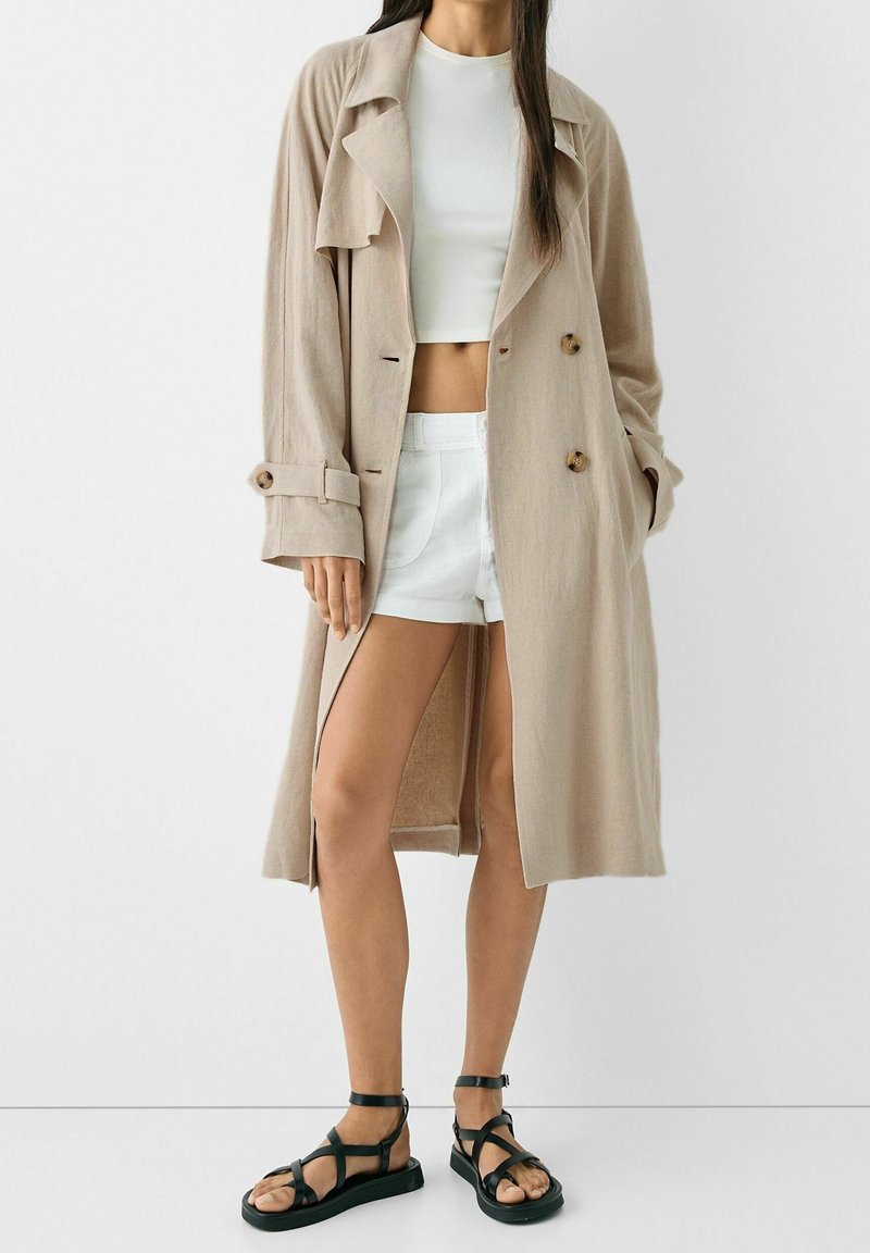 Beżowy długi płaszcz z kołnierzem, z detalami zapinanymi na guziki oraz bocznymi rozcięciami, w połączeniu z białym topem crop i białymi szortami, oraz czarnymi sandałami na paski.