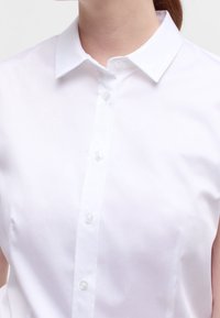 Chemise à col blanc avec une texture lisse, dotée d'une fermeture à boutons sur le devant et de détails de couture subtils, conçue pour une silhouette ajustée.