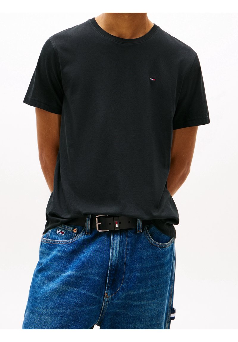 Camiseta de algodón negra con cuello redondo, mangas cortas y un pequeño logo en el pecho, combinada con pantalones de mezclilla azules y un cinturón negro.