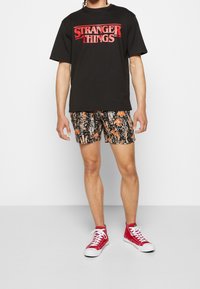 Hombre vestido con una camiseta negra de "Stranger Things", pantalones cortos negros con un patrón floral y zapatillas rojas de caña alta, de pie contra un fondo blanco.