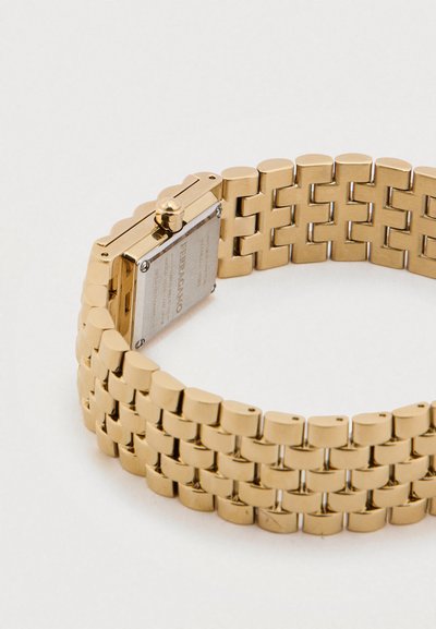 Zlaté kovové hodinky s tkaným remienkom a obdĺžnikovým puzdrom. Majú striebornú zadnú časť a výraznú korunku na nastavenie času.