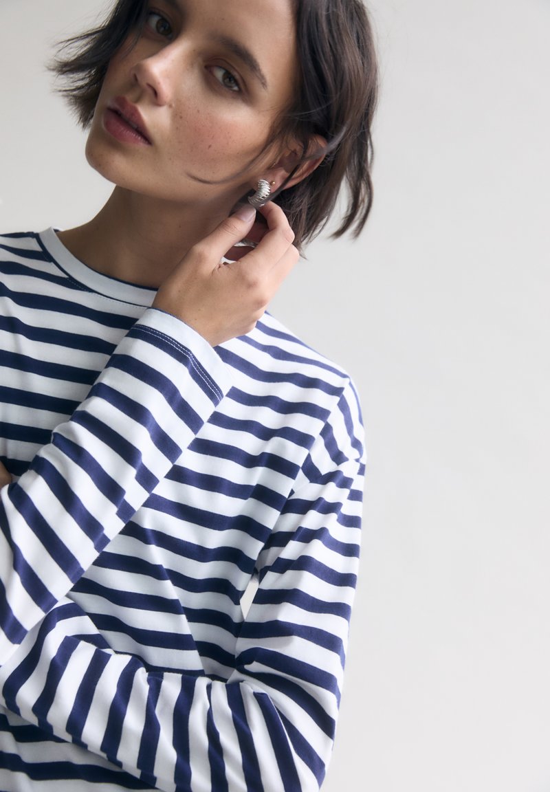 Camicia a maniche lunghe a righe blu navy e bianche con colletto rotondo, tessuto morbido e cuciture visibili, abbinata a un orecchino a cerchio argentato.