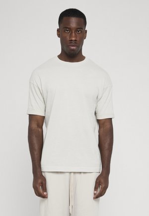 Mand, der står oprejst iført en ensfarvet hvid t-shirt med korte ærmer og lyse teksturerede bukser mod en neutral baggrund.
