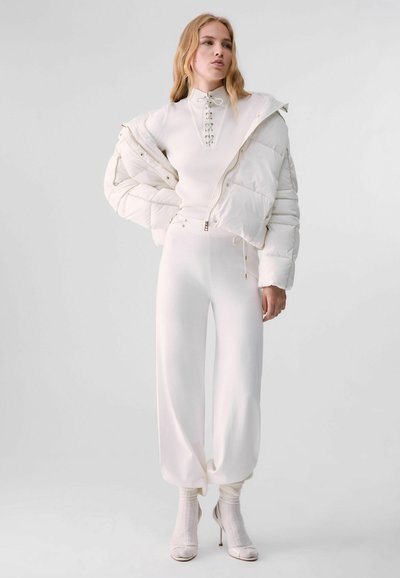 Jeune femme portant une tenue entièrement blanche comprenant une doudoune posée sur les épaules, un pantalon taille haute et des bottines à talons.