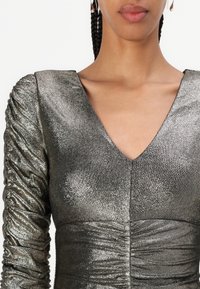 Robe argentée métallisée avec un décolleté en V profond, des manches longues froncées et un ruché à la taille, présentant un tissu texturé avec une finition brillante.