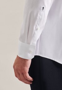 Camicia bianca a maniche lunghe con polsini abbottonati, caratterizzata da un dettaglio di rosa ricamata nera sulla manica e una texture di cotone morbido.