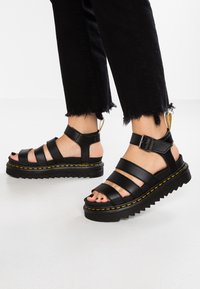Sandales en cuir noir avec trois larges brides, support de cheville à boucle, épaisse semelle à platforme avec bord dentelé et détails de couture jaune.