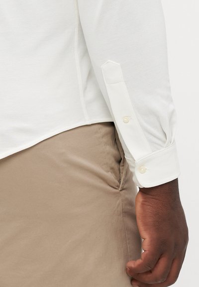 Chemise blanche à manches longues avec des poignets boutonnés, fabriquée en tissu texturé, associée à un pantalon beige clair. La chemise présente un ourlet légèrement arrondi.