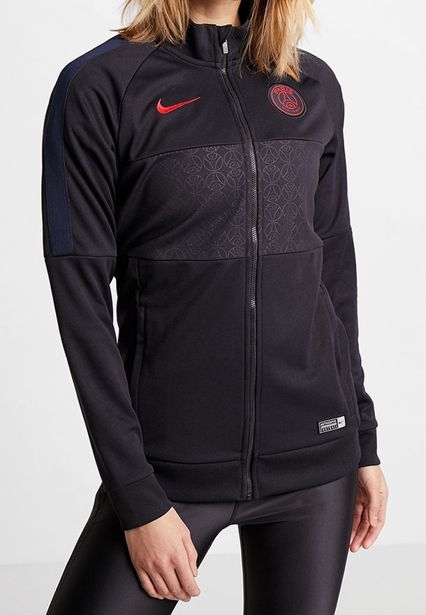 Giacca sportiva nera con zip, con loghi rossi di Nike e Paris Saint-Germain, indossata con leggings neri lucidi, su una persona con pelle chiara.