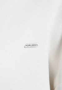 Hvid sweatshirt lavet af blødt stof, med et lille sølv mærke med teksten "SMILODOX." Designet er simpelt uden yderligere mønstre.