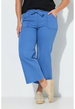 Frau trägt blaue, weit geschnittene Hosen mit Fronttaschen und gebundener Taille, beige legere Schnürschuhe, steht vor einer weißen Wand.