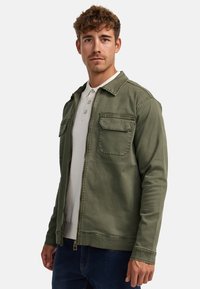 Grüne Zip-Jacke mit zwei Vordertaschen, leichtem Stoff, einem Kragen und einer glatten Textur, getragen über einem hellgrauen Polohemd.