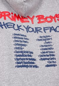 Sudadera gris con texto en negrita rojo y azul. El texto incluye "GRIMEY BOYS" y "CHECK YOUR FACE", con una lista de títulos en azul. Textura de tela suave.