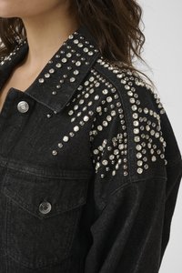 Svart denimjakke med sølvnagler og rhinestones som dekorerer kragen, skuldrene og den øvre ermet, båret av en person med bølgete brunt hår.